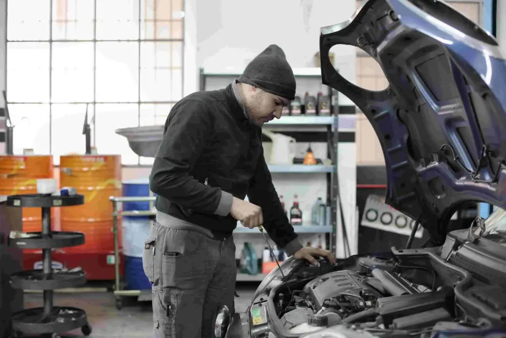 A mechanic wearing a beanie and dark clothing is inspecting the engine of a car with its hood open inside a garage. Shelves with various bottles and tools are visible in the background, highlighting the meticulous care often required to ensure vehicles meet insurance standards.