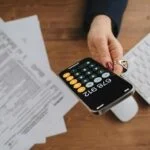 A person holds a smartphone displaying a calculator with the number 679,012. The hand has red nails and a ring. On the desk, there are insurance documents, a pen, and a white keyboard.