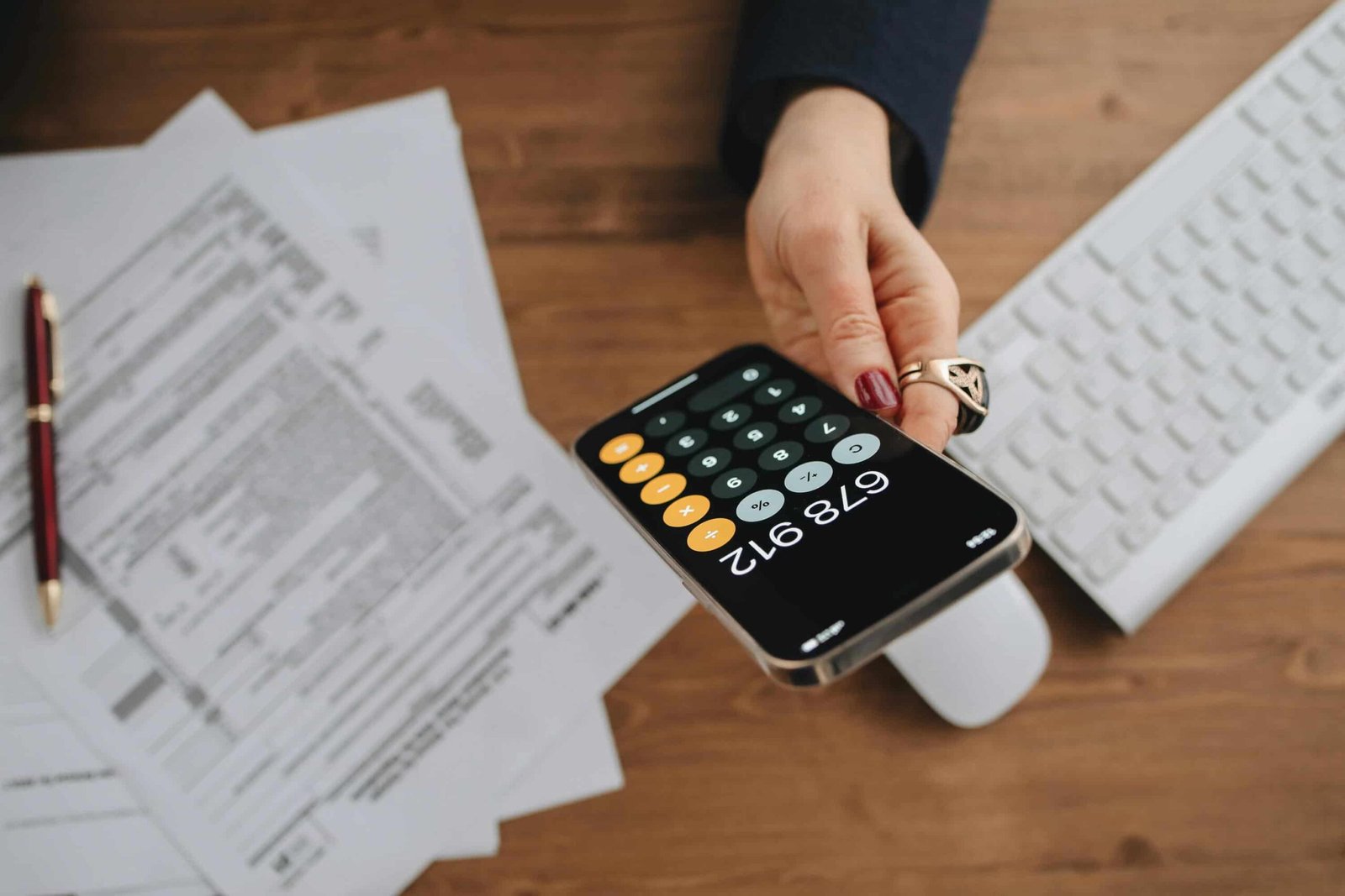 A person holds a smartphone displaying a calculator with the number 679,012. The hand has red nails and a ring. On the desk, there are insurance documents, a pen, and a white keyboard.