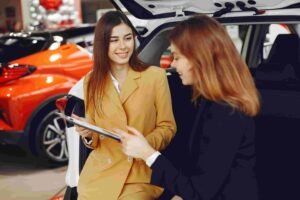 Two women are sitting in the open trunk of a car, with other vehicles visible in the background. One woman in a yellow coat is holding a clipboard, possibly related to insurance, while the other in a black coat is engaged in conversation. Both are smiling and seemingly discussing something.