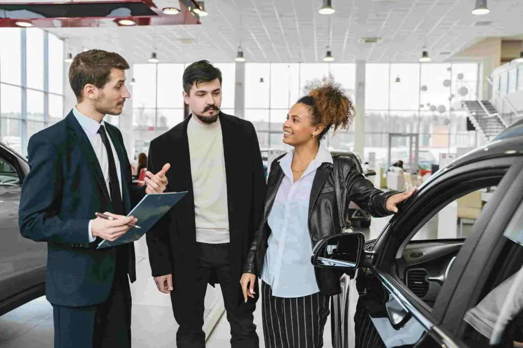 Three people are in a car showroom. A salesperson in a suit is talking while holding a clipboard, discussing insurance options. A man and a woman stand next to him, listening attentively. The woman leans on a black car, smiling, while the man looks at the salesperson.