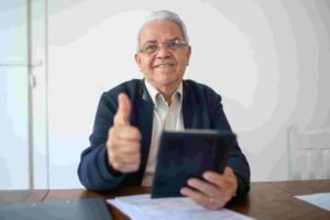 An elderly man with white hair and glasses smiles warmly while holding a tablet in one hand and giving a thumbs-up with the other. He sits at a wooden table with some insurance documents on it, against a plain white background.