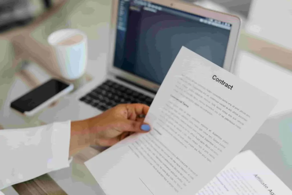 A person holding a printed insurance contract, with a blurred background showing a laptop, coffee mug, and smartphone on a desk. The person's nails are painted light blue. The laptop screen displays a program with code.