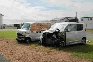 Two damaged vans parked on grass, one silver with a crumpled front and covered partially with plastic, the other white with makeshift cardboard and tape repairs on the front and side. Residential houses are visible in the background under a cloudy sky—a scene reflecting neglected insurance claims.