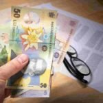 A hand holds several Romanian leu banknotes of different denominations. The background shows a pair of eyeglasses, partially visible insurance documents, and part of a calculator on a wooden surface.