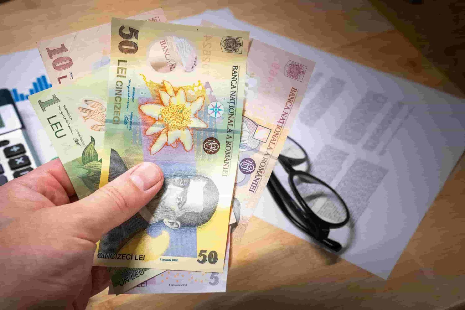 A hand holds several Romanian leu banknotes of different denominations. The background shows a pair of eyeglasses, partially visible insurance documents, and part of a calculator on a wooden surface.