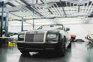 A sleek, black luxury car with a distinctive grille and hood ornament is parked inside a modern, well-lit garage with geometric lighting fixtures on the ceiling. Various workstations and car parts can be seen in the background, ensuring all repairs are done perfectly before your insurance claim.