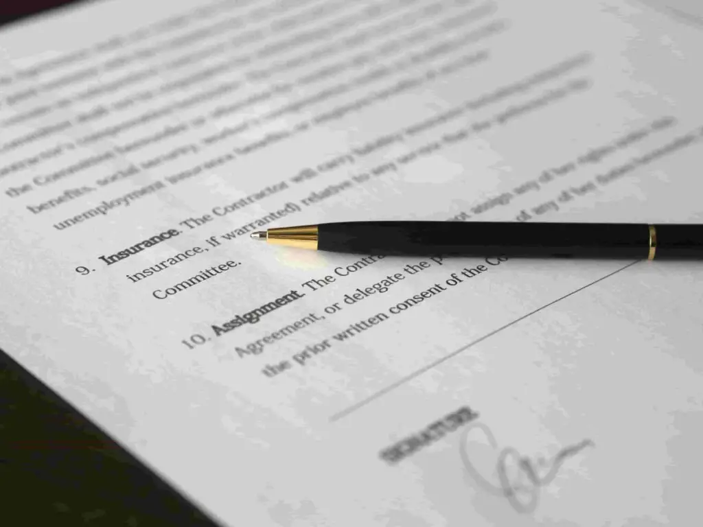 A black and gold pen rests on a document containing typed text. Only part of the document is visible, showing section headings such as "Insurance" and "Assignment." Near the bottom, there's an illegible signature on the signature line.