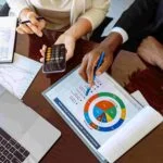 Two people are sitting at a table engaged in a discussion. One person is holding a calculator, and the other is pointing at a colorful insurance chart on a document. A laptop, notebook, and some charts are also on the table.