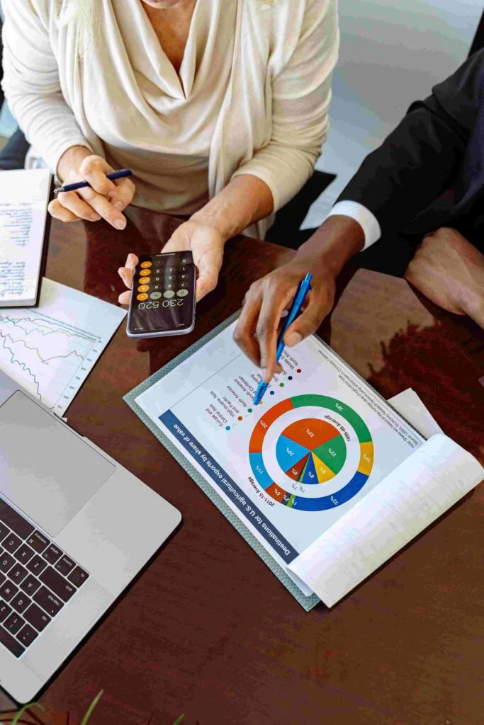 Two people are sitting at a table engaged in a discussion. One person is holding a calculator, and the other is pointing at a colorful insurance chart on a document. A laptop, notebook, and some charts are also on the table.