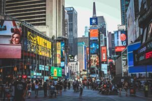 A bustling city scene captures Times Square in New York City, filled with vibrant billboards, advertisements for everything from Broadway shows to insurance, and electronic screens. Crowds of people walk through the streets, and towering skyscrapers rise in the background on a bright day.