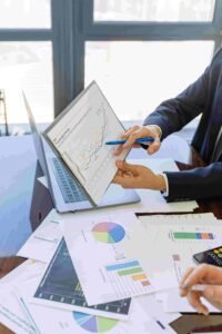 Two business professionals are analyzing financial data using a laptop and various paper reports, focusing on insurance trends. One person is pointing to a graph on the laptop screen with a blue pen. The table is cluttered with pie charts, bar graphs, and other documents.