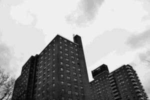 Black-and-white photo of two tall brick apartment buildings viewed from below. The structures, covered by many windows and some balconies, exude a vintage charm. Under an overcast sky, leafless tree branches peek into the frame. A striking image that subtly underscores the need for comprehensive insurance coverage.