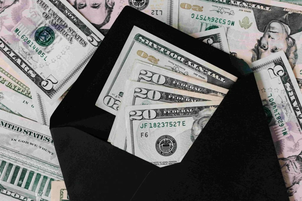 A black envelope partially opened, showing several US dollar bills, including twenties and fives, surrounded by more scattered US currency—perhaps a payout from an insurance claim.