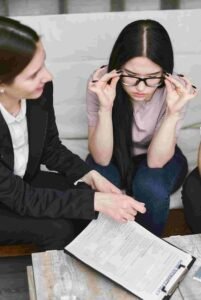 Two individuals are sitting on a couch looking at an insurance document on a clipboard. One person is holding her glasses while attentively reading the document. The other individual, wearing a blazer, is pointing at the document, likely explaining its contents.