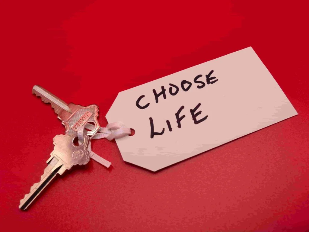 A pair of silver keys are attached to a white tag on a red background. The tag has the handwritten message "CHOOSE LIFE" in bold, black letters.