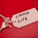 A pair of silver keys are attached to a white tag on a red background. The tag has the handwritten message "CHOOSE LIFE" in bold, black letters.