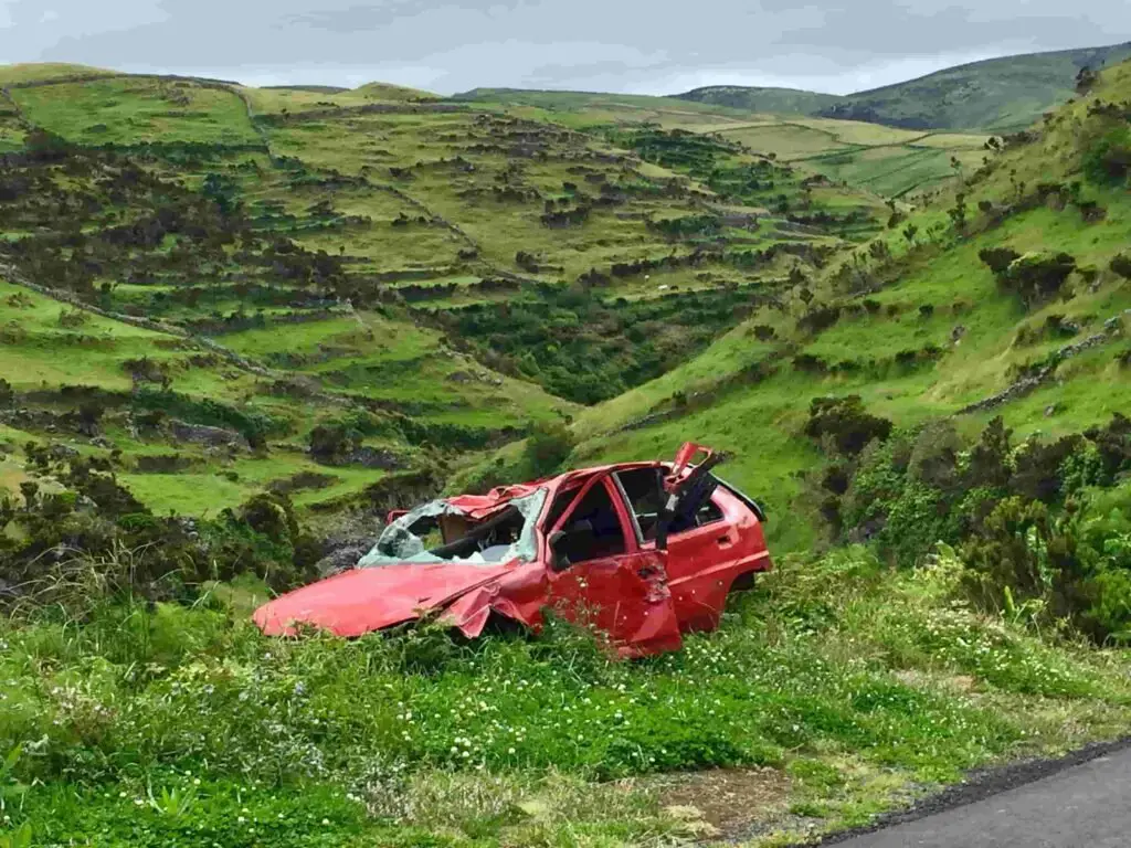 A red car with significant damage sits abandoned in a grassy, green hilly landscape, a testament to the importance of insurance. The car's roof and windows are crushed, suggesting it rolled over. The surrounding scenery is lush with vegetation and rolling hills under an overcast sky.