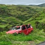 A red car with significant damage sits abandoned in a grassy, green hilly landscape, a testament to the importance of insurance. The car's roof and windows are crushed, suggesting it rolled over. The surrounding scenery is lush with vegetation and rolling hills under an overcast sky.