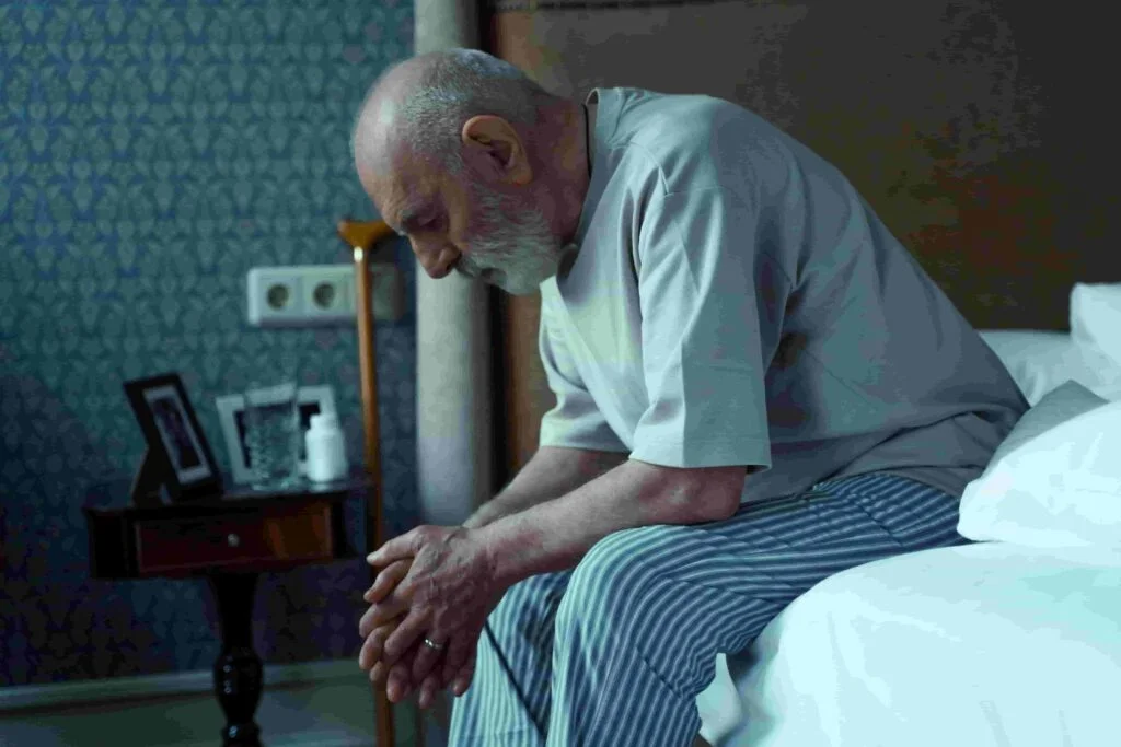 An elderly man wearing a light grey shirt and striped pajama pants sits on the edge of a bed with his head bowed, appearing deep in thought. A walking cane is propped against a nightstand that holds framed photos and a white bottle. The room is dimly lit.