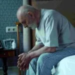 An elderly man wearing a light grey shirt and striped pajama pants sits on the edge of a bed with his head bowed, appearing deep in thought. A walking cane is propped against a nightstand that holds framed photos and a white bottle. The room is dimly lit.