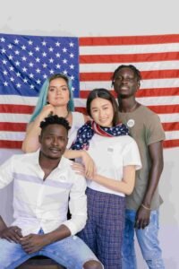 Four people stand and sit in front of an American flag. Two stand, and two sit, posing for the camera. One person wears an American flag scarf. They all have relaxed, confident expressions and a "Vote" sticker on their clothing.