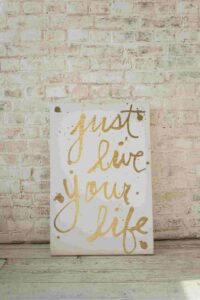 A white poster with a golden cursive inscription that reads "just live your life" is placed against a whitewashed brick wall. The poster has splashes of gold paint around the text. The scene has a rustic, artistic vibe.