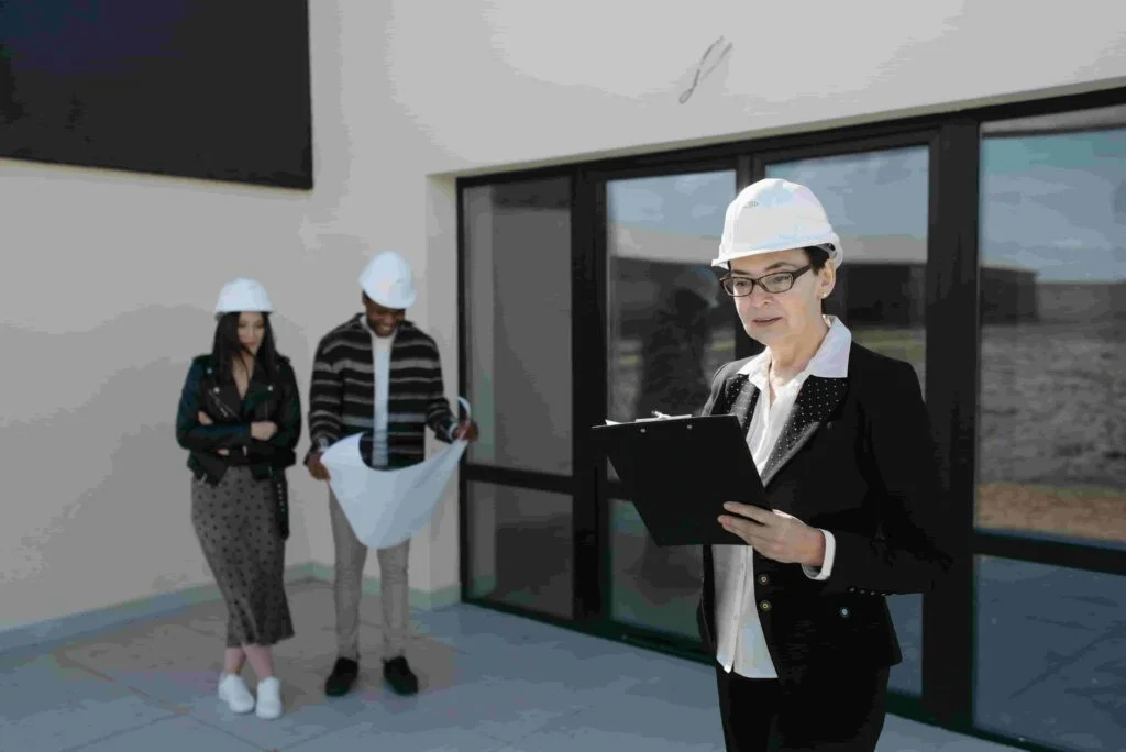 A woman in a construction helmet reads from a clipboard outside a property. In the background, a man and woman, also wearing helmets, examine blueprints. They're all dressed in business attire.