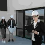 A woman in a construction helmet reads from a clipboard outside a property. In the background, a man and woman, also wearing helmets, examine blueprints. They're all dressed in business attire.