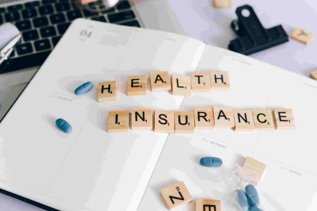 An open planner on a desk displays the words "Health Insurance" spelled with wooden letter tiles. Nearby are blue and white capsules, a pen, a laptop keyboard, and a black clipboard—reminders of the essential role insurance plays in safeguarding health.