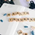 An open planner on a desk displays the words "Health Insurance" spelled with wooden letter tiles. Nearby are blue and white capsules, a pen, a laptop keyboard, and a black clipboard—reminders of the essential role insurance plays in safeguarding health.