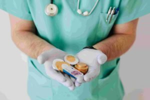 A person wearing teal medical scrubs and white gloves holds a pile of assorted cryptocurrencies, represented by physical coins, in their cupped hands. A stethoscope is draped around their neck, hinting at a modern intersection of healthcare and insurance markets.