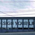 A building facade with large windows features the words "DESIGN YOUR LIFE" in bold black letters, echoing the assurance of a tailored insurance plan. The background is an abstract pattern, and power lines stretch across the sky.