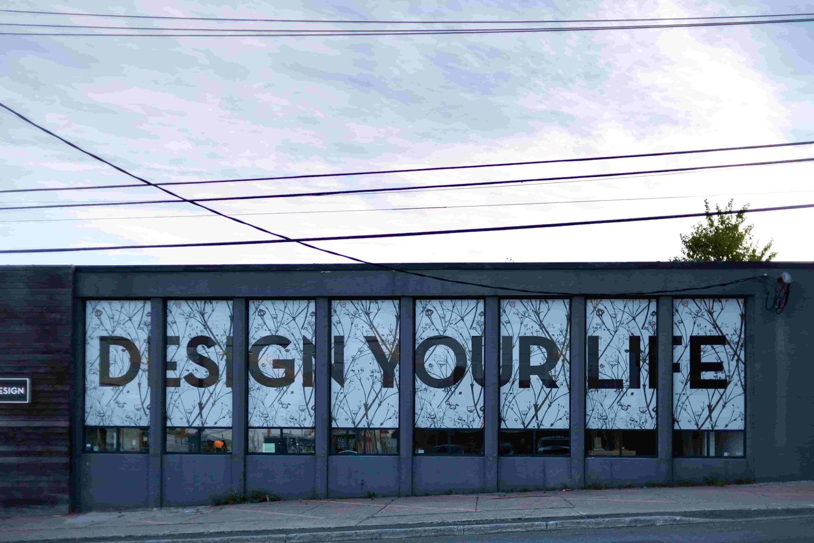 A building facade with large windows features the words "DESIGN YOUR LIFE" in bold black letters, echoing the assurance of a tailored insurance plan. The background is an abstract pattern, and power lines stretch across the sky.