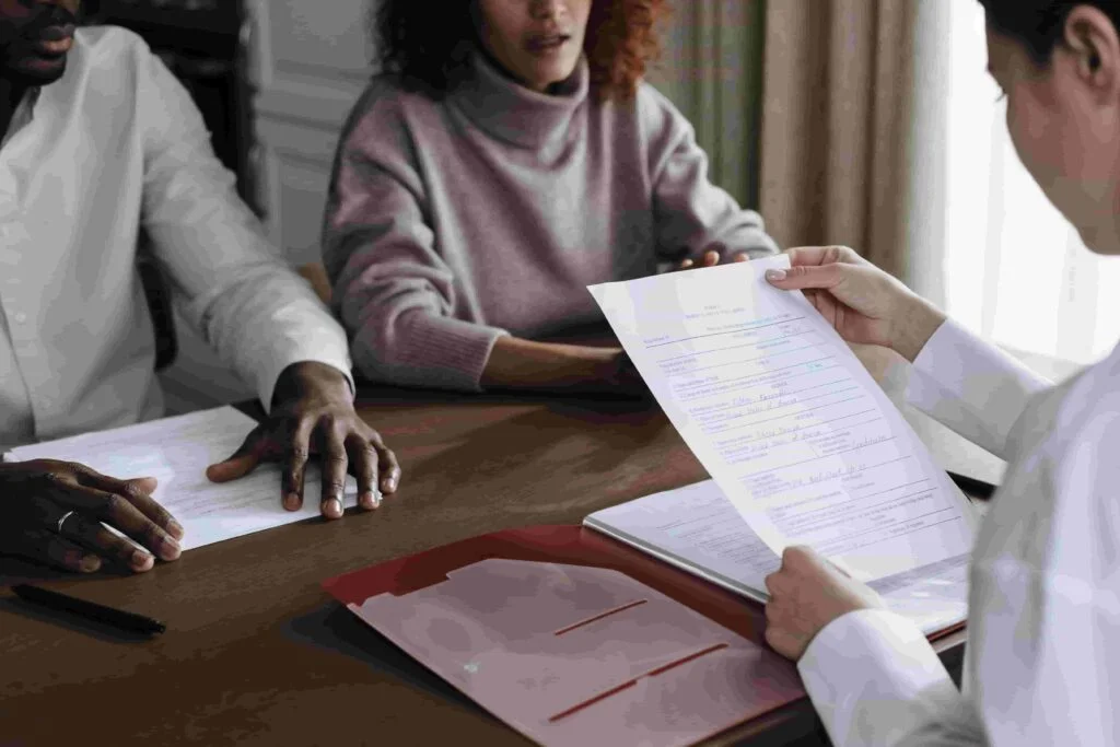 A close-up of a formal meeting. Three people are seated at a table discussing insurance matters. One holds a document folder, another holds a sheet of paper, and the third person looks on. A maroon folder is open on the table.