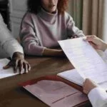 A close-up of a formal meeting. Three people are seated at a table discussing insurance matters. One holds a document folder, another holds a sheet of paper, and the third person looks on. A maroon folder is open on the table.