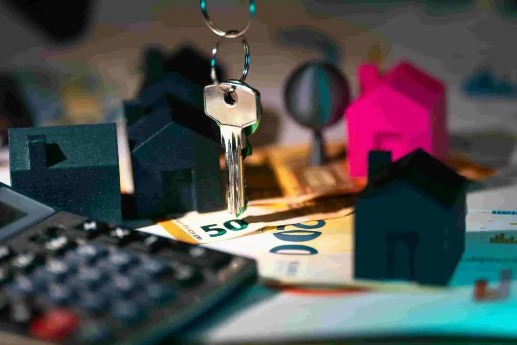 A close-up of a key hanging above euro banknotes, with small black and pink house figurines in the background. Nearby, a calculator and an insurance document symbolize real estate investment or home buying protection.