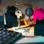 A close-up of a key hanging above euro banknotes, with small black and pink house figurines in the background. Nearby, a calculator and an insurance document symbolize real estate investment or home buying protection.