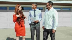 Three people are standing outside in front of a house with a garage. A woman in a red dress is animatedly discussing insurance options with two men, one in a shirt and tie, holding a policy paper, and the other in a white shirt and glasses.