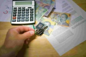 A hand holds keys over Romanian currency and documents on a wooden table, hinting at real estate or financial activities. Nearby, a calculator partially covered by banknotes and insurance forms suggests a blend of calculations, paperwork, and currency matters.