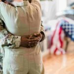 A person in military uniform embraces another indoors, with an American flag partially visible in the background, symbolizing the care and dedication often mirrored in healthcare.