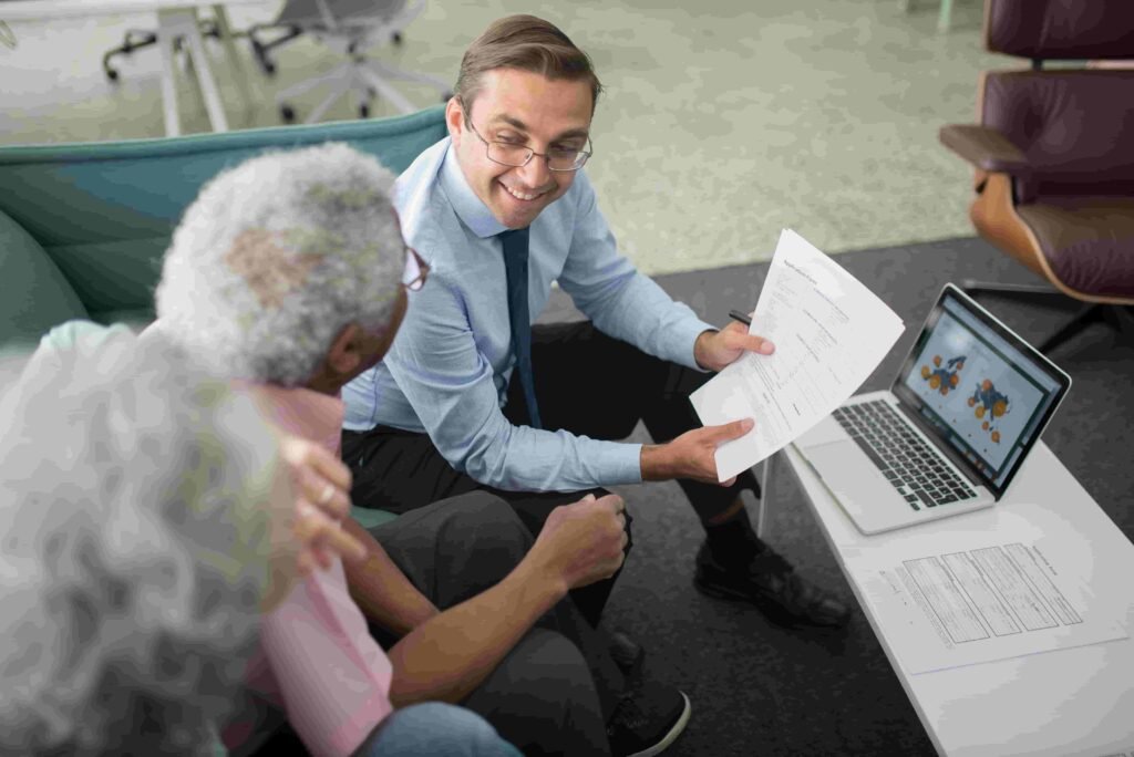 A man in a light blue shirt and tie holds an insurance document, carefully explaining it to an elderly couple seated across from him. A laptop displaying a map is open on the table in front of them. The setting appears to be a modern office with sleek furniture.