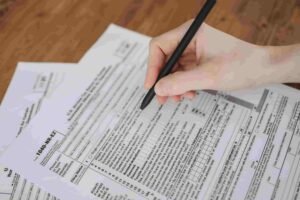 A hand holding a black pen is meticulously filling out a 1040 tax form on a wooden table, with several pages detailing deductions and insurance information. The form, partially completed, features small printed text and boxes awaiting essential details.