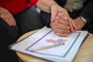 Two people hold hands above a document with charts, placed on a table. A pen rests on the document, hinting at an insurance discussion. The scene suggests comfort and support during the meeting.