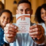 A person holds a pamphlet titled Voluntary Life Insurance Policy with a family symbol and the words Extra protection for your family. In the background, a smiling family of four is seated together.