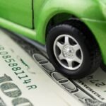 A toy green car sits atop a pile of hundred-dollar bills, symbolizing money and financial transactions related to vehicles.
